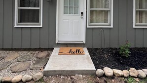 A single-story house with a white front door, two windows on either side, and a 'hello' mat at the entrance.