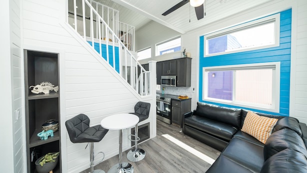 A compact living space with a staircase leading to an upper level, a kitchen area, a black leather sofa, and a small dining table with two chairs.