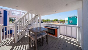 A wooden deck with a white railing, a wooden table with two chairs, and a built-in fireplace.
