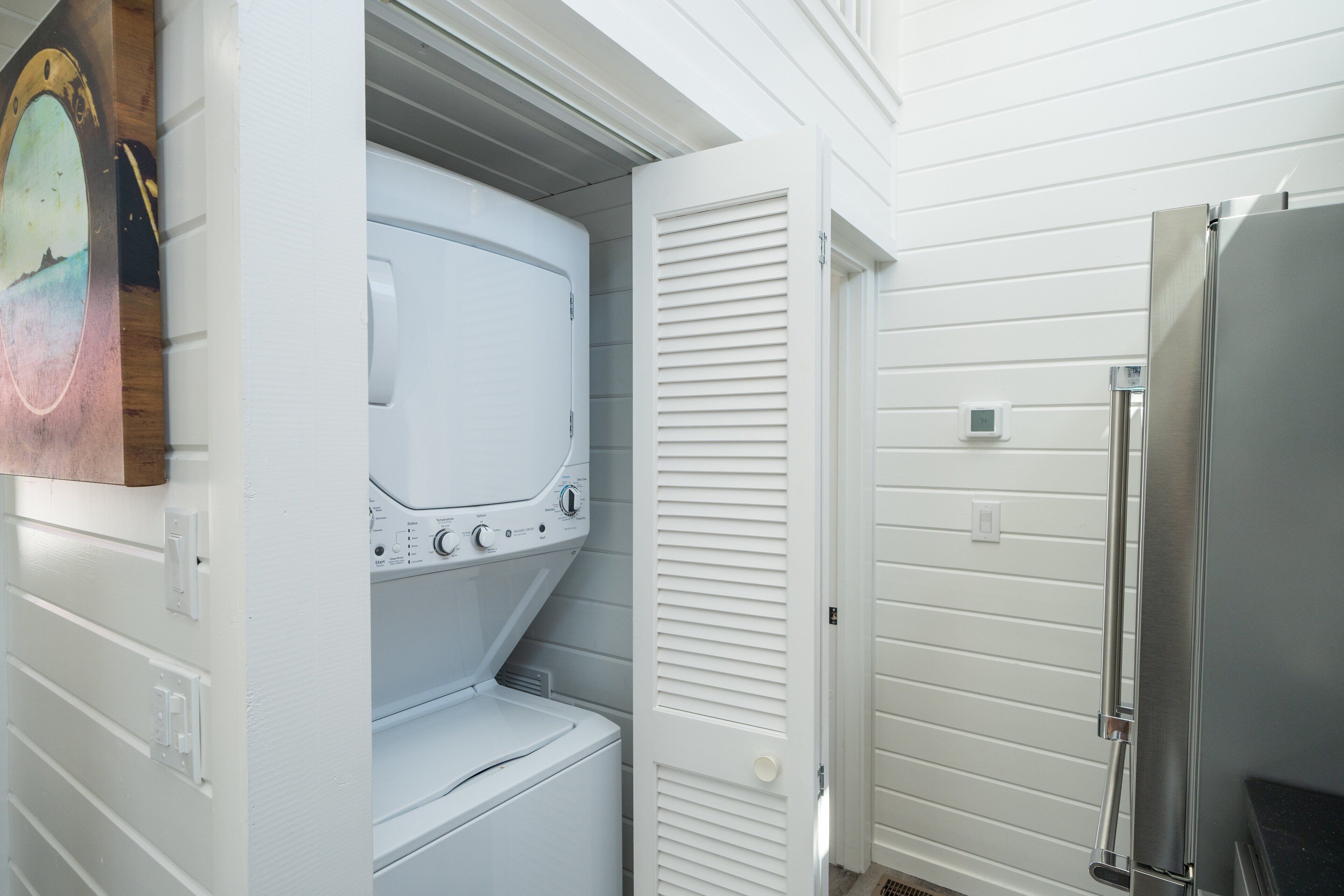 Un cuarto de lavandería con lavadora y secadora apiladas, una puerta con celosías y un refrigerador de acero inoxidable.