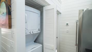 Un cuarto de lavandería con lavadora y secadora apiladas, una puerta con celosías y un refrigerador de acero inoxidable.