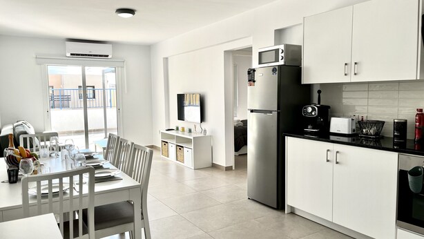 Una cocina moderna con armarios blancos, un microondas integrado y un refrigerador. Hay un comedor con una mesa y sillas, y un televisor montado en la pared.