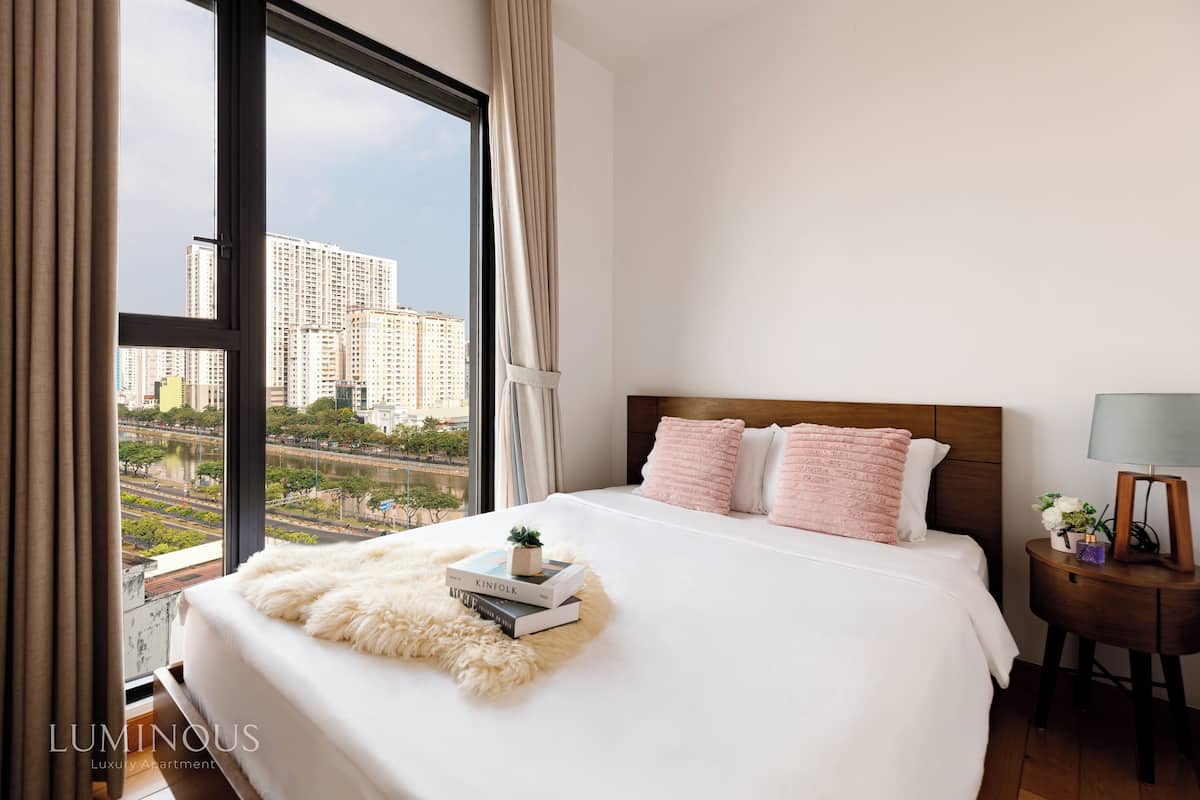 Un dormitorio con un ventanal grande, una cama con ropa de cama blanca y vistas a edificios altos.