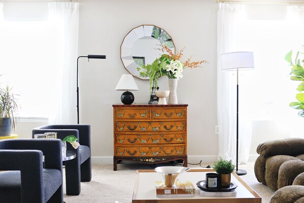 A living room with a blue sofa, a wooden dresser with floral carvings, a round mirror, and a coffee table with a vase of flowers.