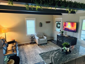 Un salon avec un canapé en cuir, un fauteuil blanc, un téléviseur, une cheminée et un plafond orné de plantes vertes.