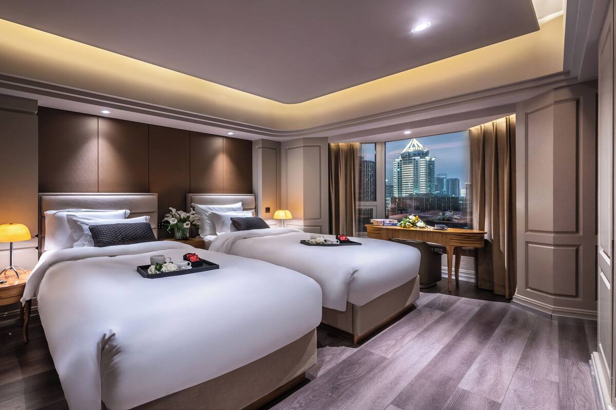 Habitación de hotel con dos camas, un escritorio, una ventana con vistas a la ciudad y una mesita de noche con lámpara.