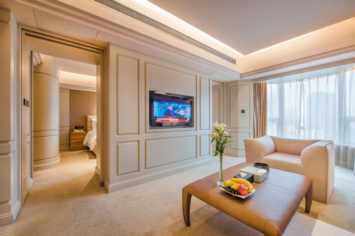 Habitación de hotel con televisión en la pared, sofá, mesa de centro con frutas y un jarrón con flores.