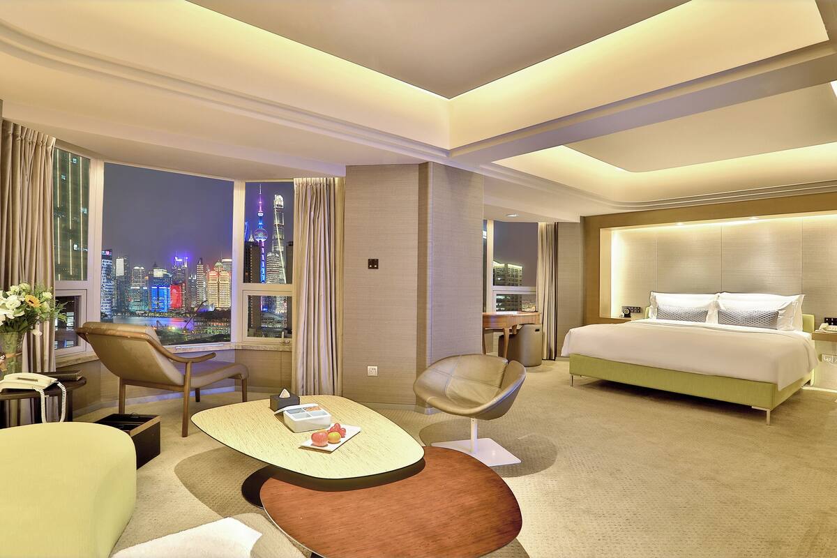 Habitación de hotel moderna con vista a la ciudad, una cama grande, un escritorio con una silla y una mesita con un florero y fruta.