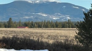 A mountain with snow-capped peaks, a field with sparse vegetation, and a small red-roofed building in the foreground. - Modern Tiny House in the Woods (Athol)