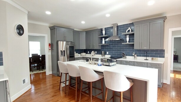 A modern kitchen with grey cabinets, a large island with bar stools, and a stainless steel refrigerator.