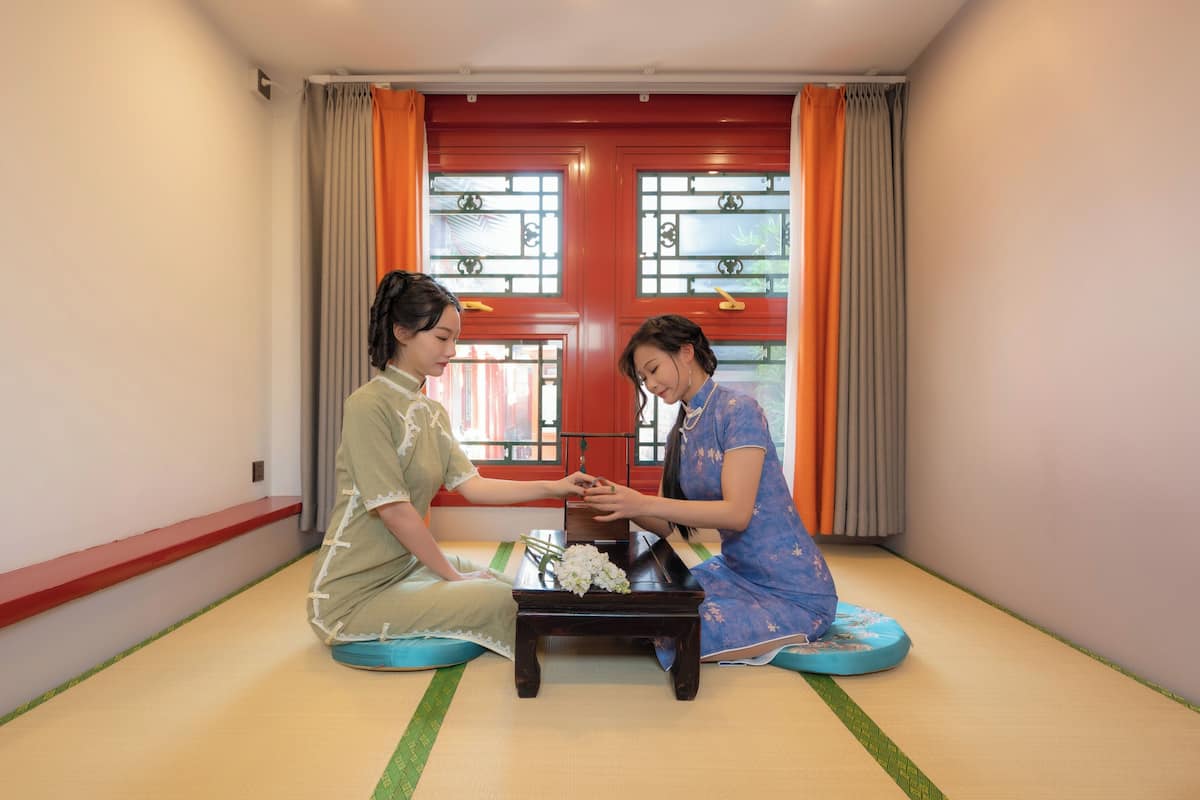 Two individuals seated on tatami mats in a room with a traditional red door and windows with decorative grilles.