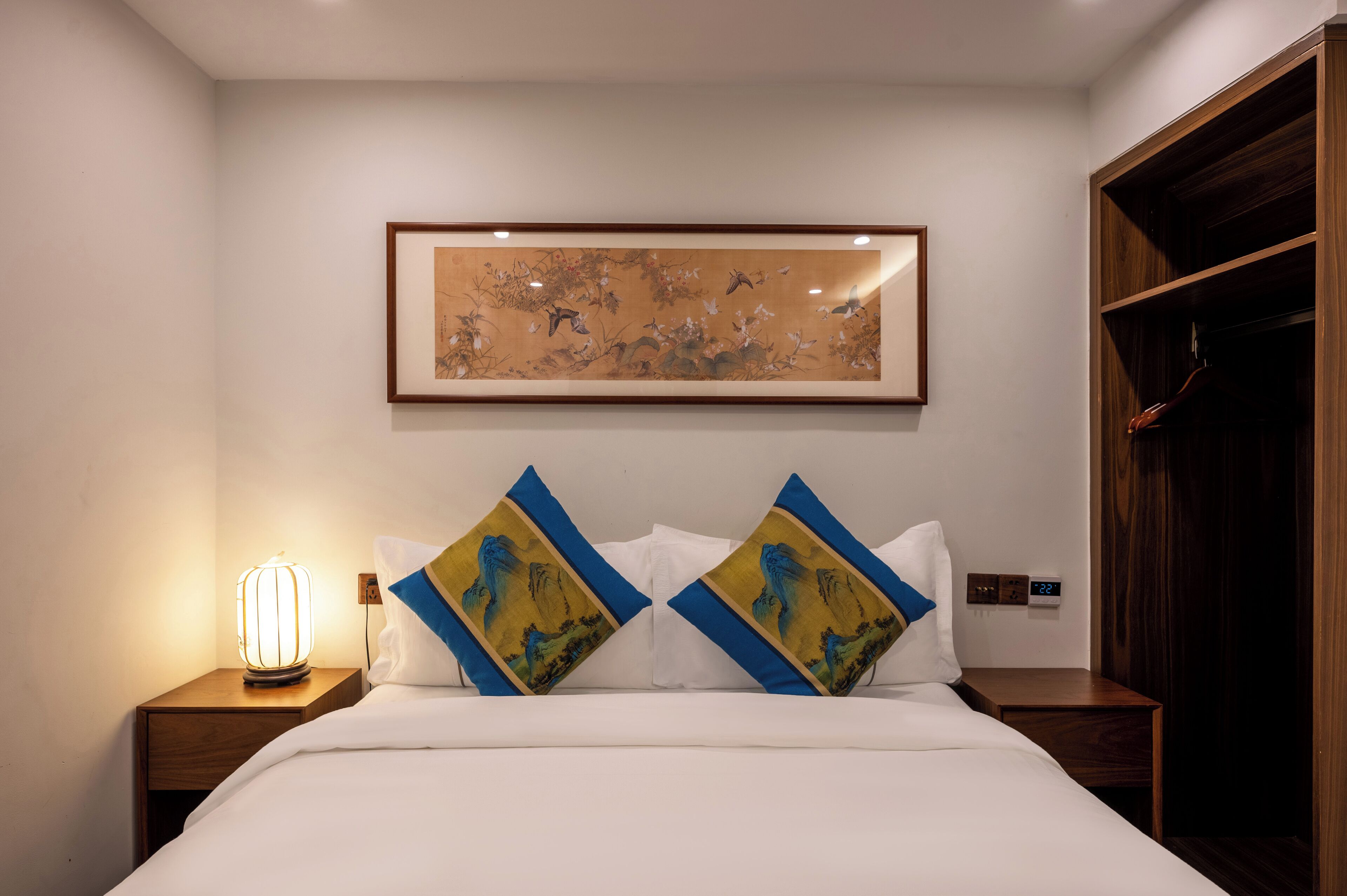 A hotel room with a bed, two pillows with a blue and yellow design, a wall-mounted artwork, a bedside lamp, and a wooden wardrobe.