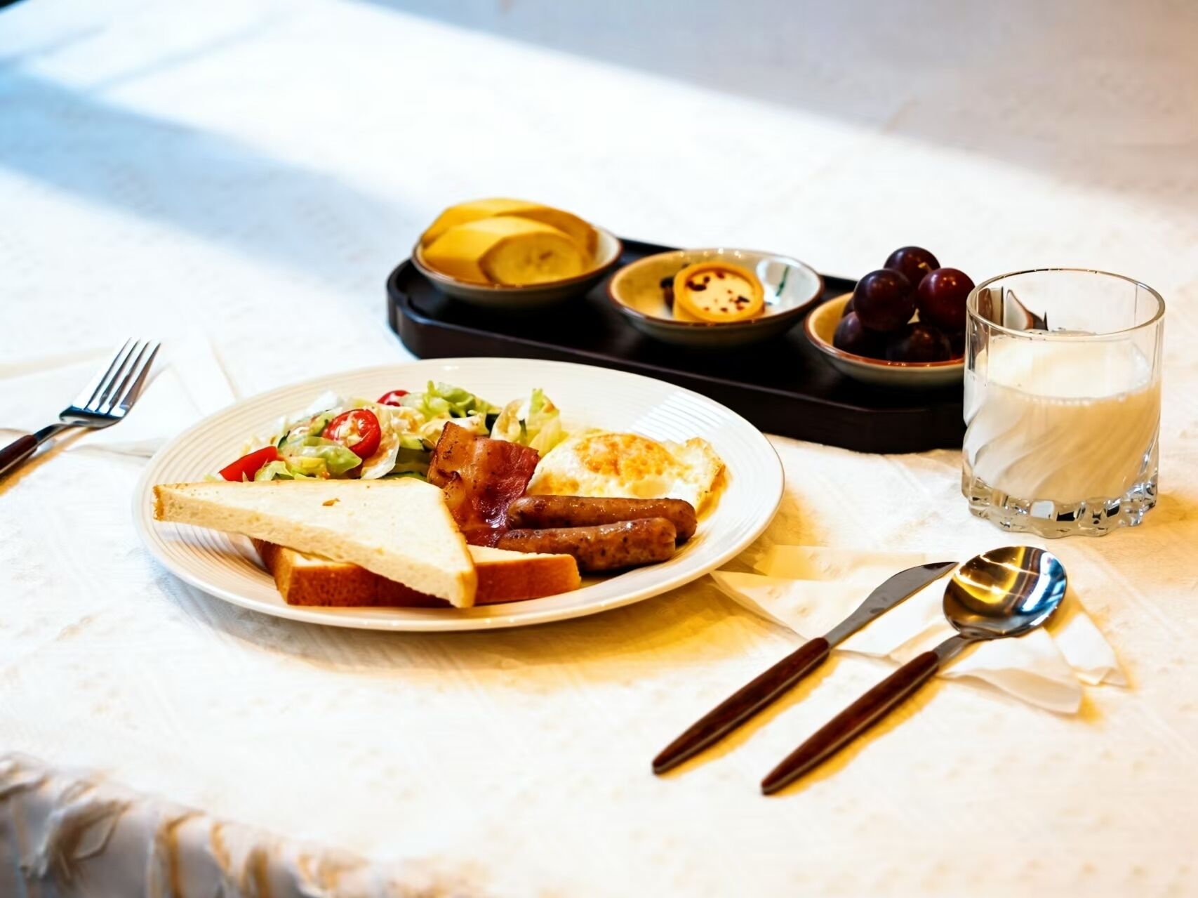 A breakfast plate with toast, bacon, sausage, and salad, a glass of milk, and a tray with a bowl of fruit and a small dish of jam.