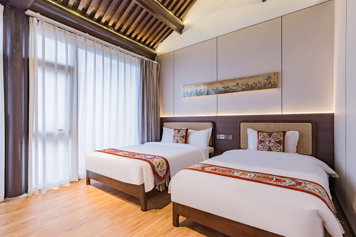 A hotel room with two beds, wooden flooring, a large window with sheer curtains, and a wall-mounted artwork.