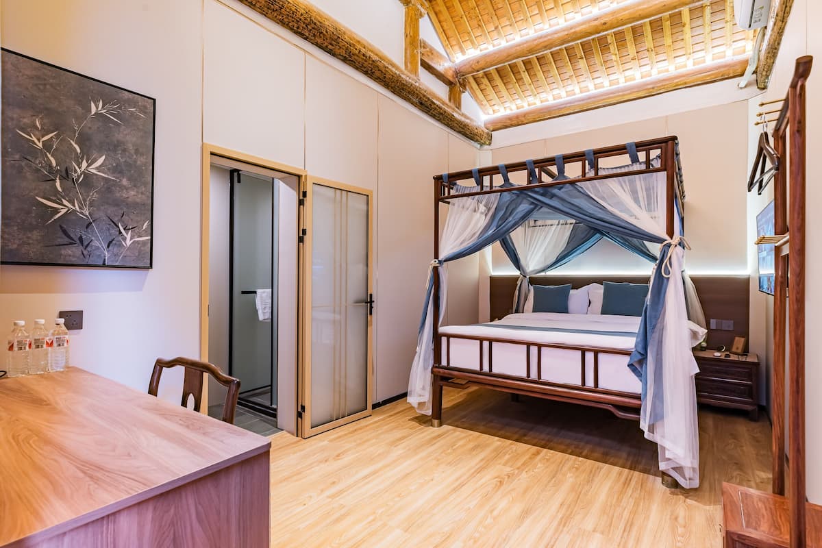 A room with a wooden floor, a bed with a canopy, a desk with chairs, a wardrobe, and a painting on the wall.