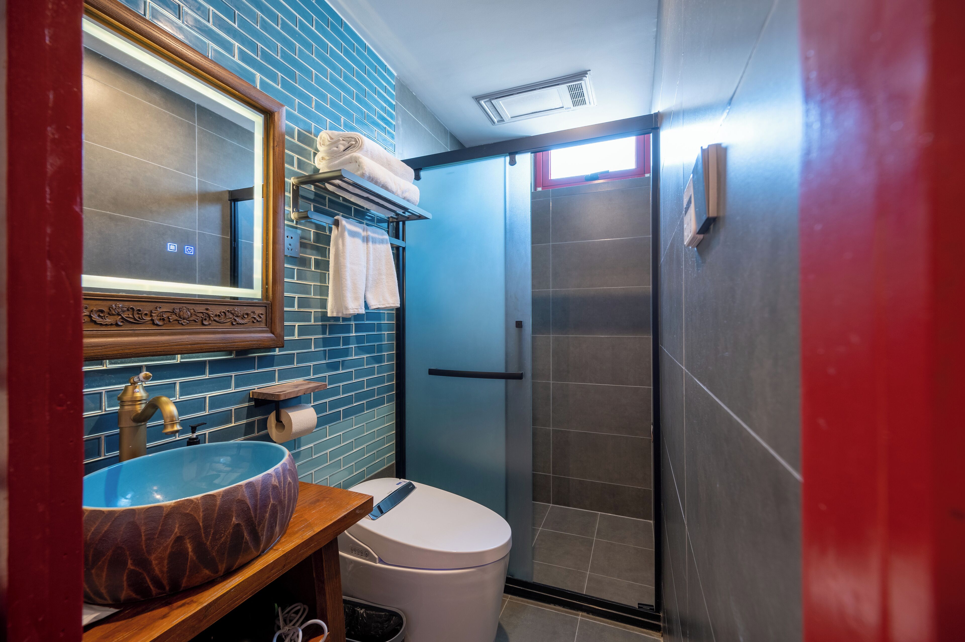 A bathroom with a blue tiled wall, a wooden vanity with a marble sink, a white toilet, and a glass shower enclosure.