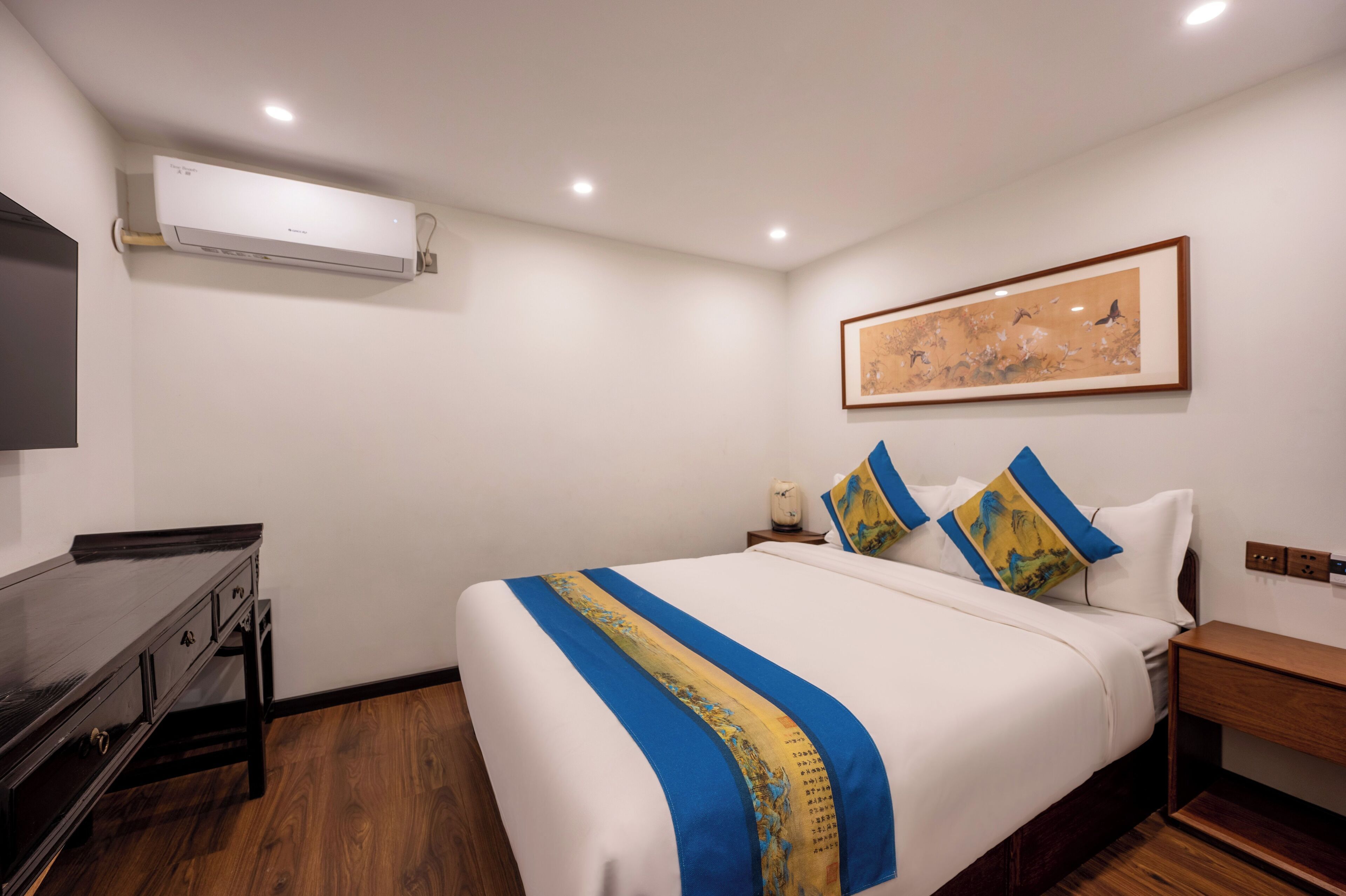 A hotel room with a bed, bedside table, nightstand, and an air conditioning unit on the wall.