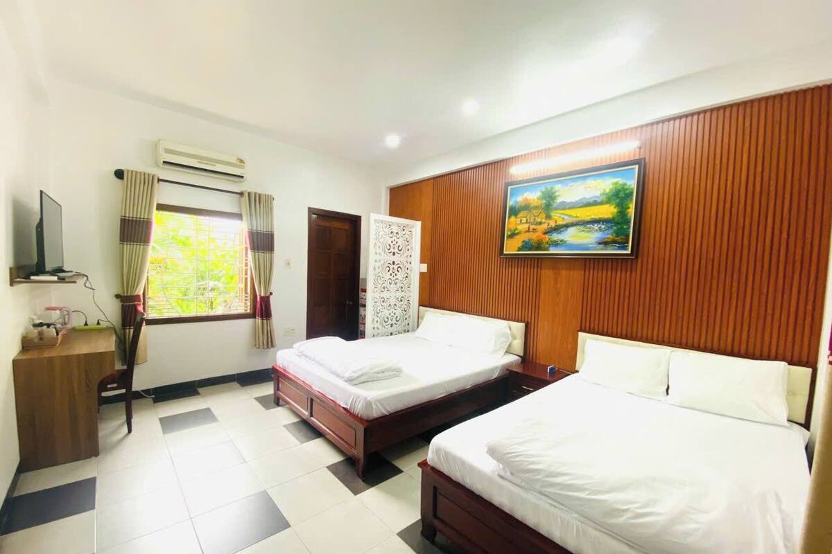A hotel room with two beds, a desk, a chair, a window with curtains, an air conditioner, and a painting on the wall.