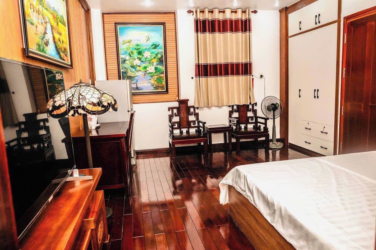 A hotel room with a wooden floor, a bed, a desk with a lamp, a chair, a fan, and a wardrobe.