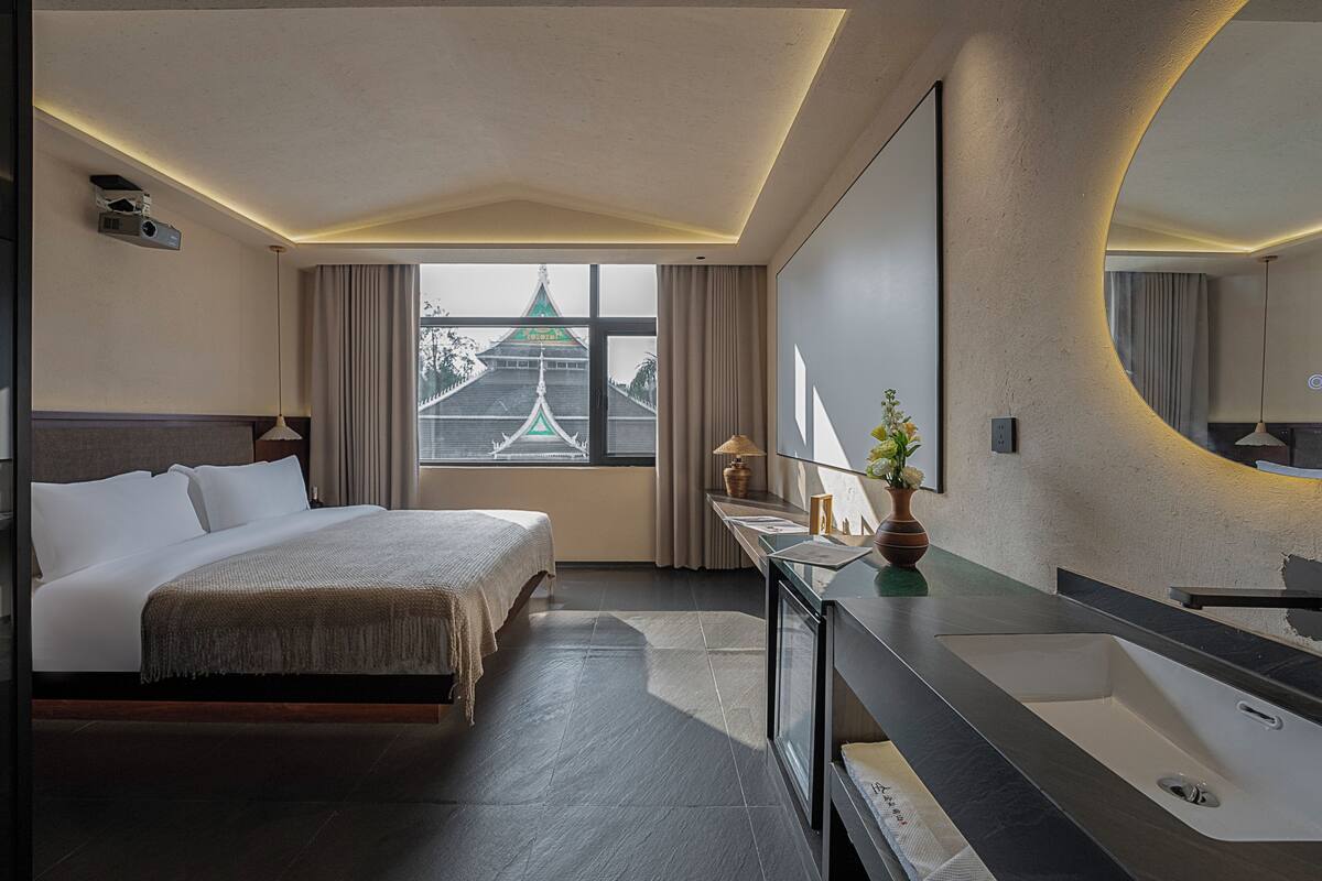 Ein modernes Hotelzimmer mit einem großen Bett, einem Badezimmer mit großem Spiegel und Waschbecken sowie Blick auf ein traditionelles Gebäude vor dem Fenster.