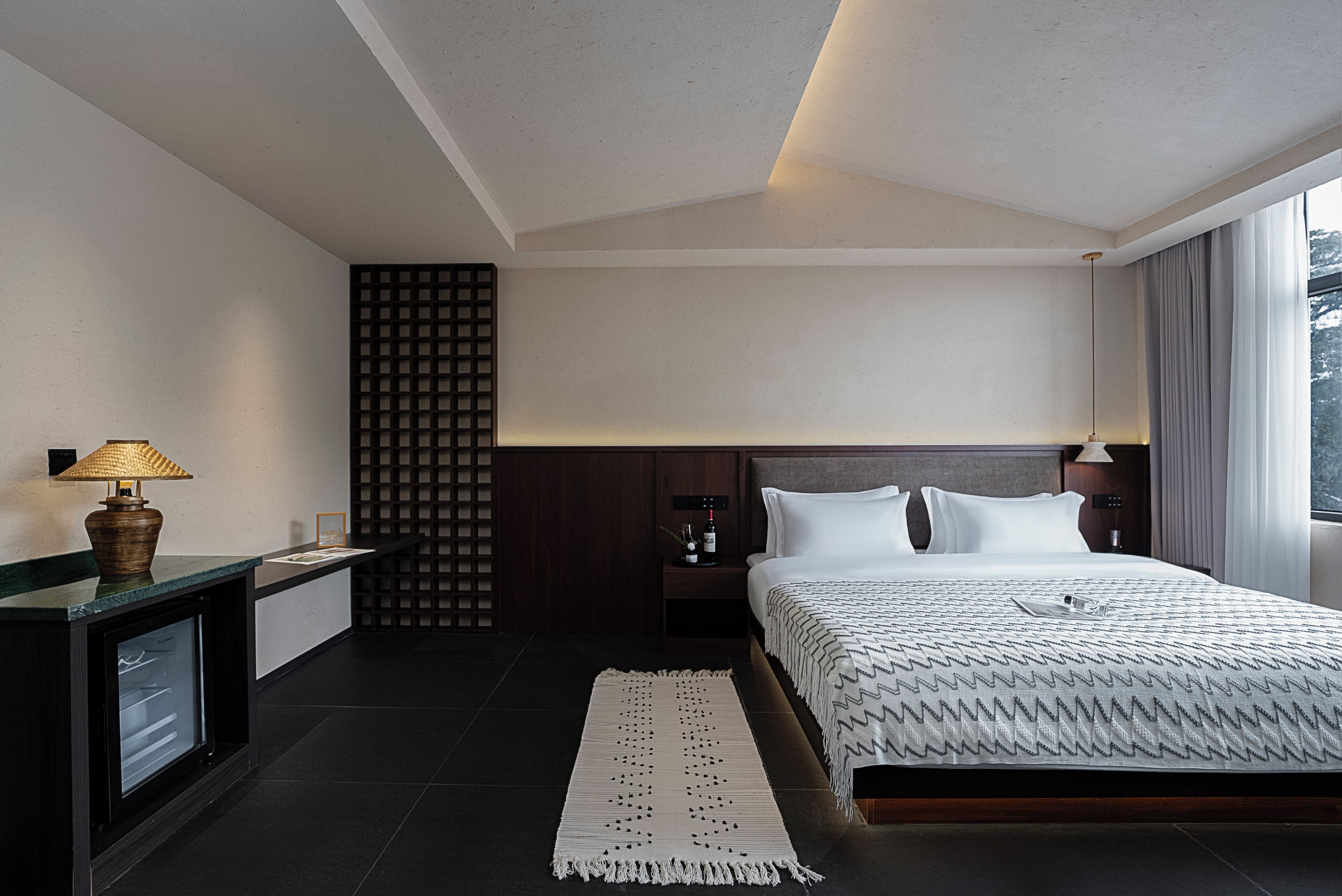 A modern hotel room with a large bed, a desk with a lamp, a minibar, and a view of the outdoors.