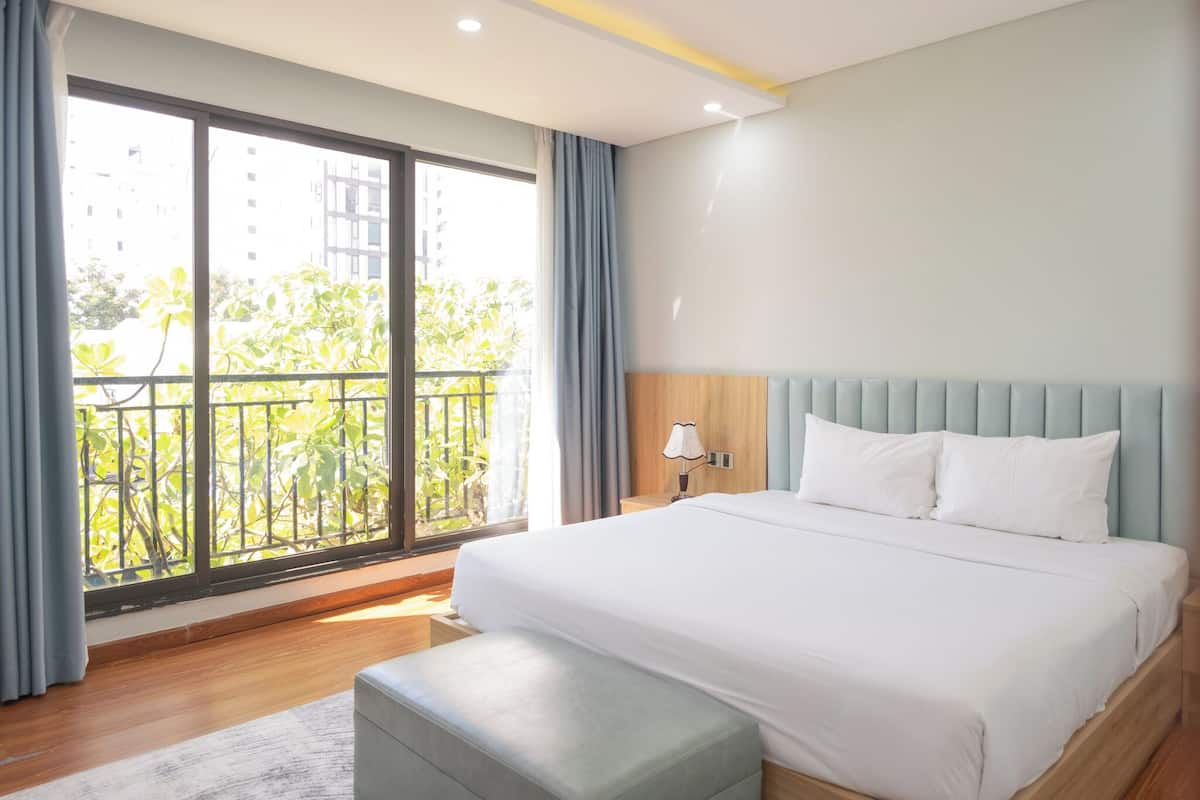 Ein modernes Hotelzimmer mit einem großen Bett, einem Nachttisch, einem Fenster mit Vorhängen und Blick auf Grünflächen und Gebäude.