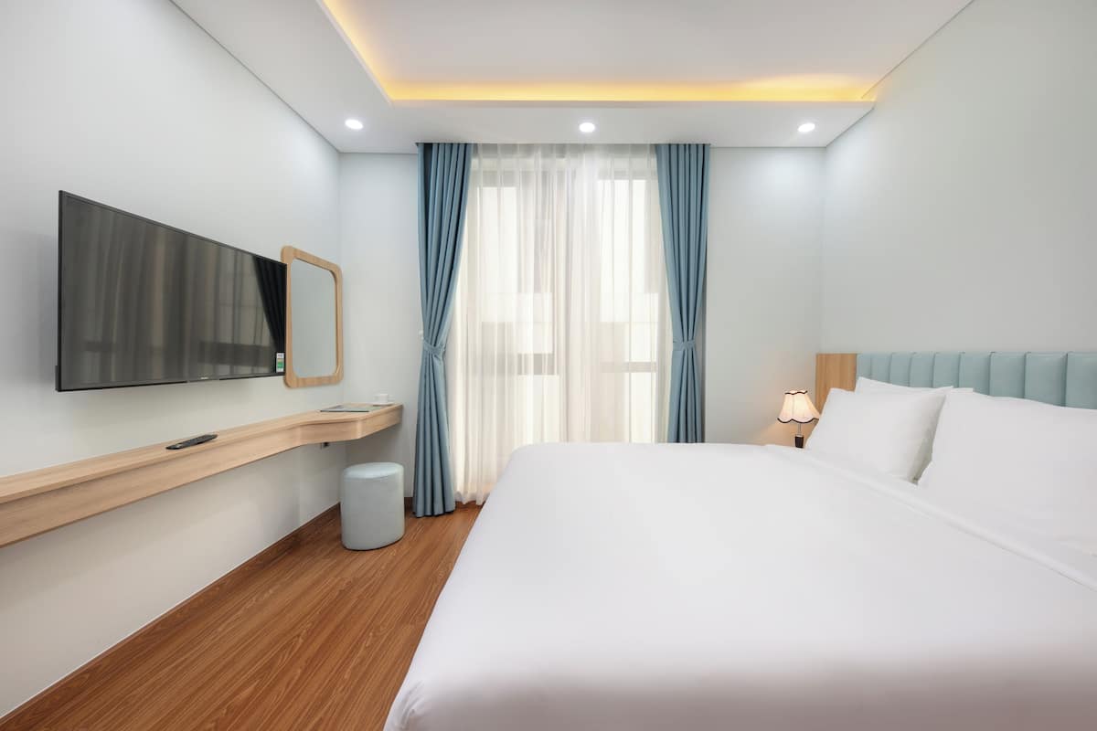 Ein modernes Hotelzimmer mit einem großen Bett, einem flachbildfernseher, der an einer hölzernen Konsole angebracht ist, einer Nachttischlampe und einem Fenster mit durchsichtigen Vorhängen.