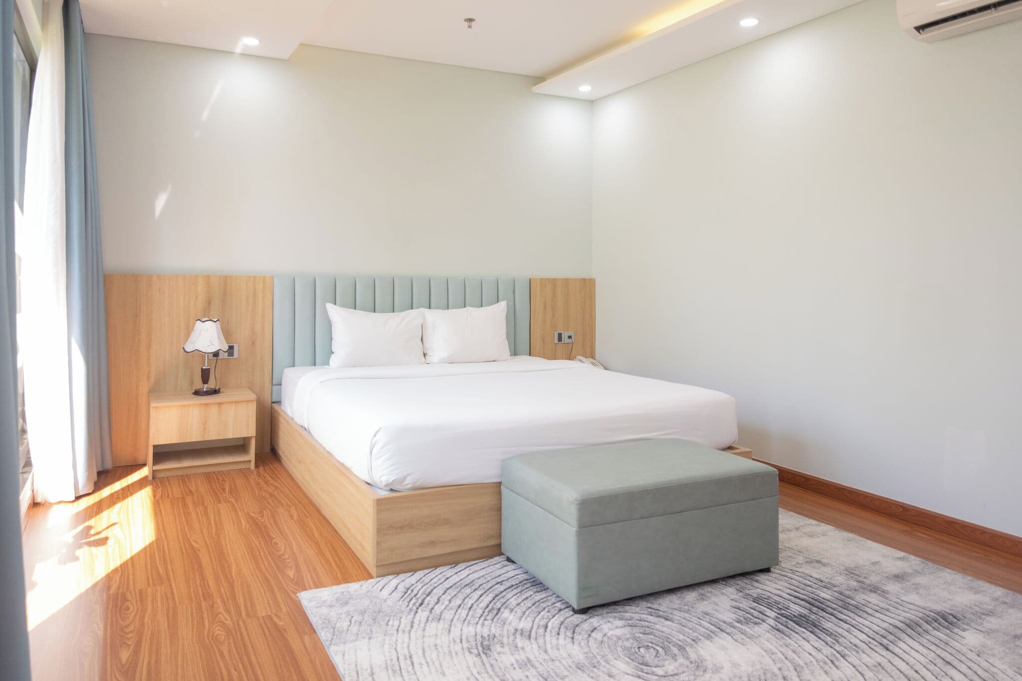 Une chambre moderne avec une tête de lit en bois, un lit recouvert de linge de maison blanc, une table de chevet avec une lampe et un pouf gris.