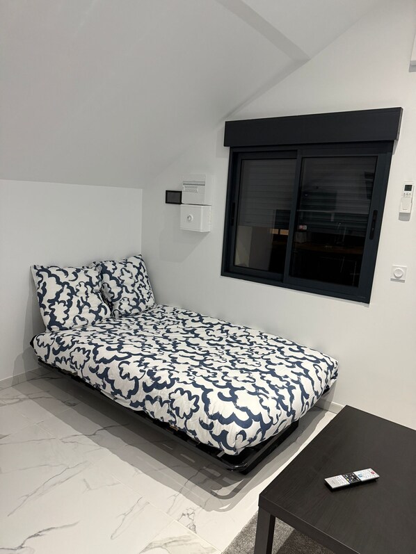 A small, neatly arranged room with a bed, a window, a wall-mounted thermostat, and a remote control on a table.