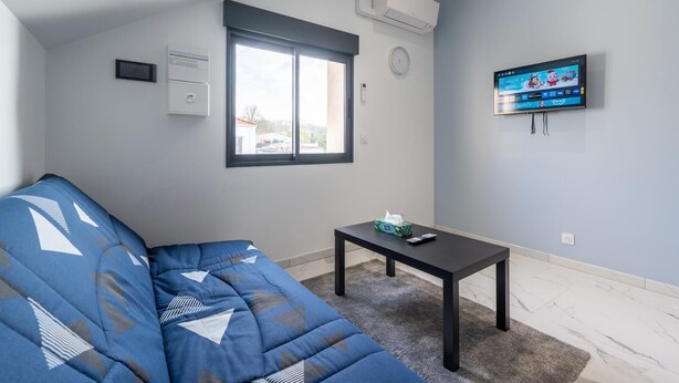 A modern living room with a blue sofa, a small table, a mounted TV, and a window with blinds. - Charming studio near Roissy CDG (Vaujours)