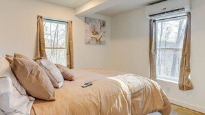 Un dormitorio con cama, dos ventanas, un equipo de aire acondicionado y un cuadro en la pared.