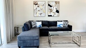A modern living room with a sectional sofa, an ottoman, and a glass coffee table. There are abstract paintings on the wall and a window with curtains.