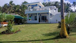 A two-story white house with a balcony, surrounded by a well-maintained lawn and palm trees. - We are directly on the beach with 50 meters of frontage and beautiful sunsets (Bobon)