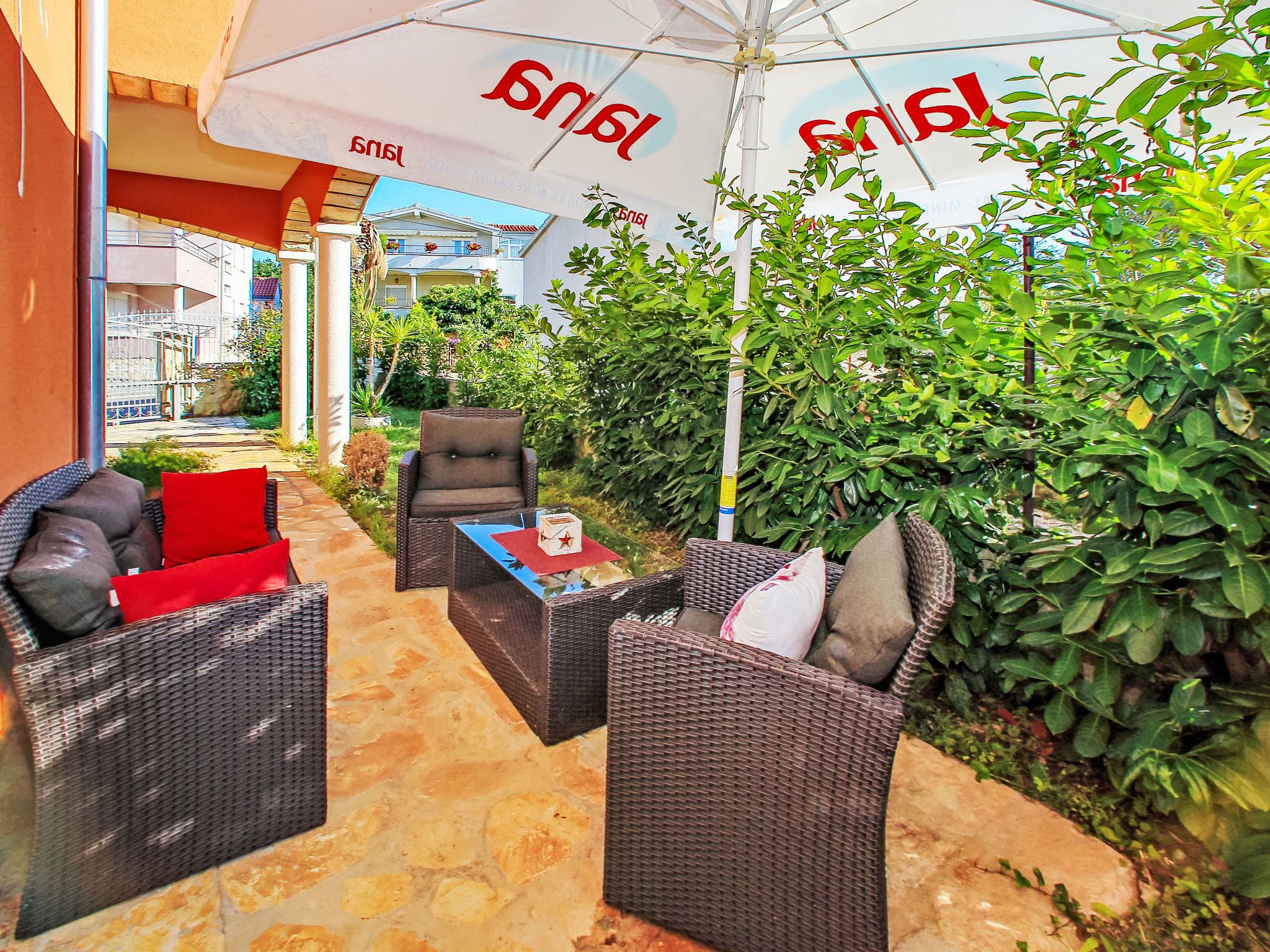 An outdoor seating area with wicker furniture and a table, surrounded by greenery and a building in the background.