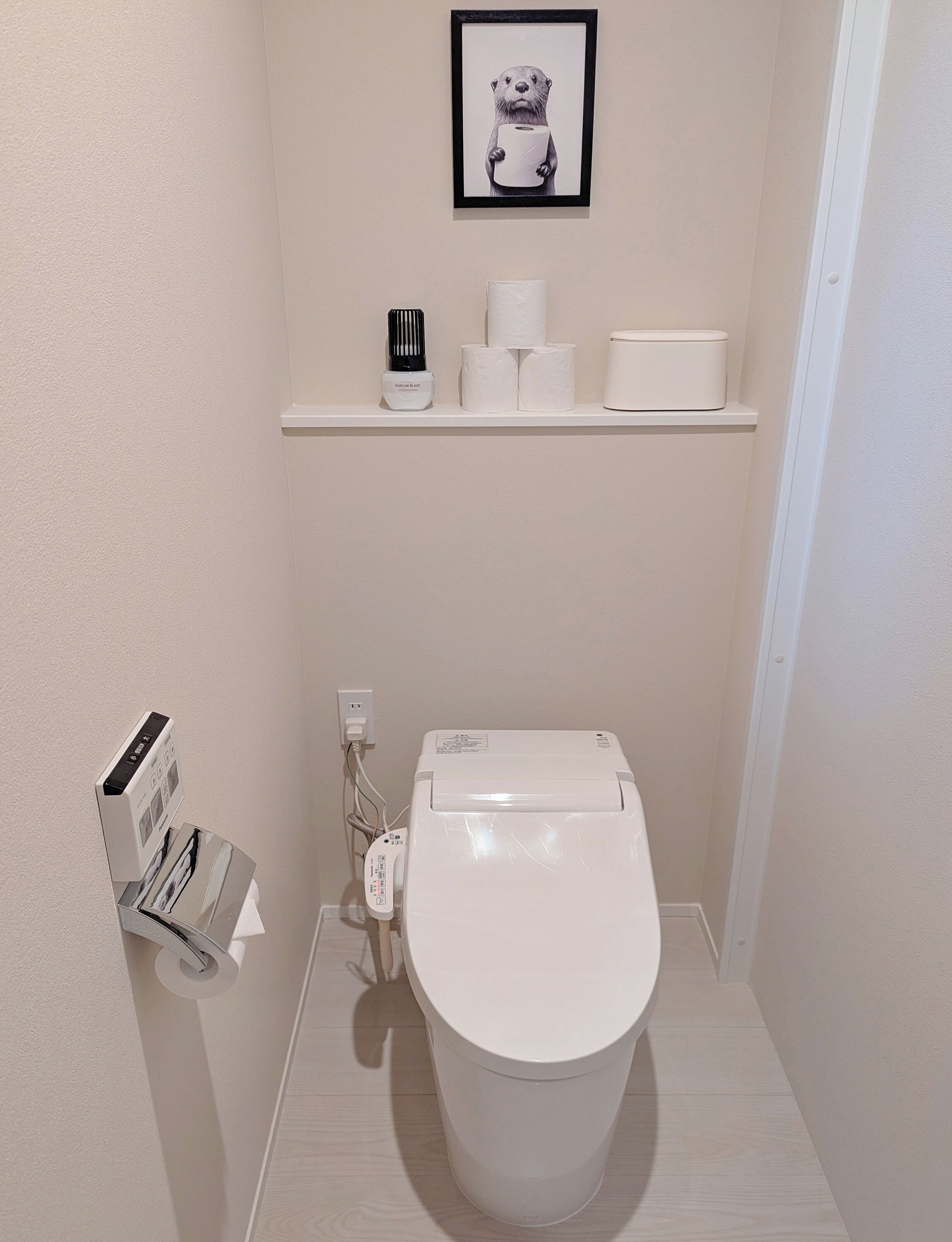 Un baño pequeño y moderno con un inodoro blanco, un estante con papel higiénico y un secador de pelo, y un cuadro en la pared.