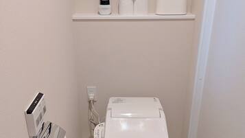 Un baño pequeño y moderno con un inodoro blanco, un estante con papel higiénico y un secador de pelo, y un cuadro en la pared.