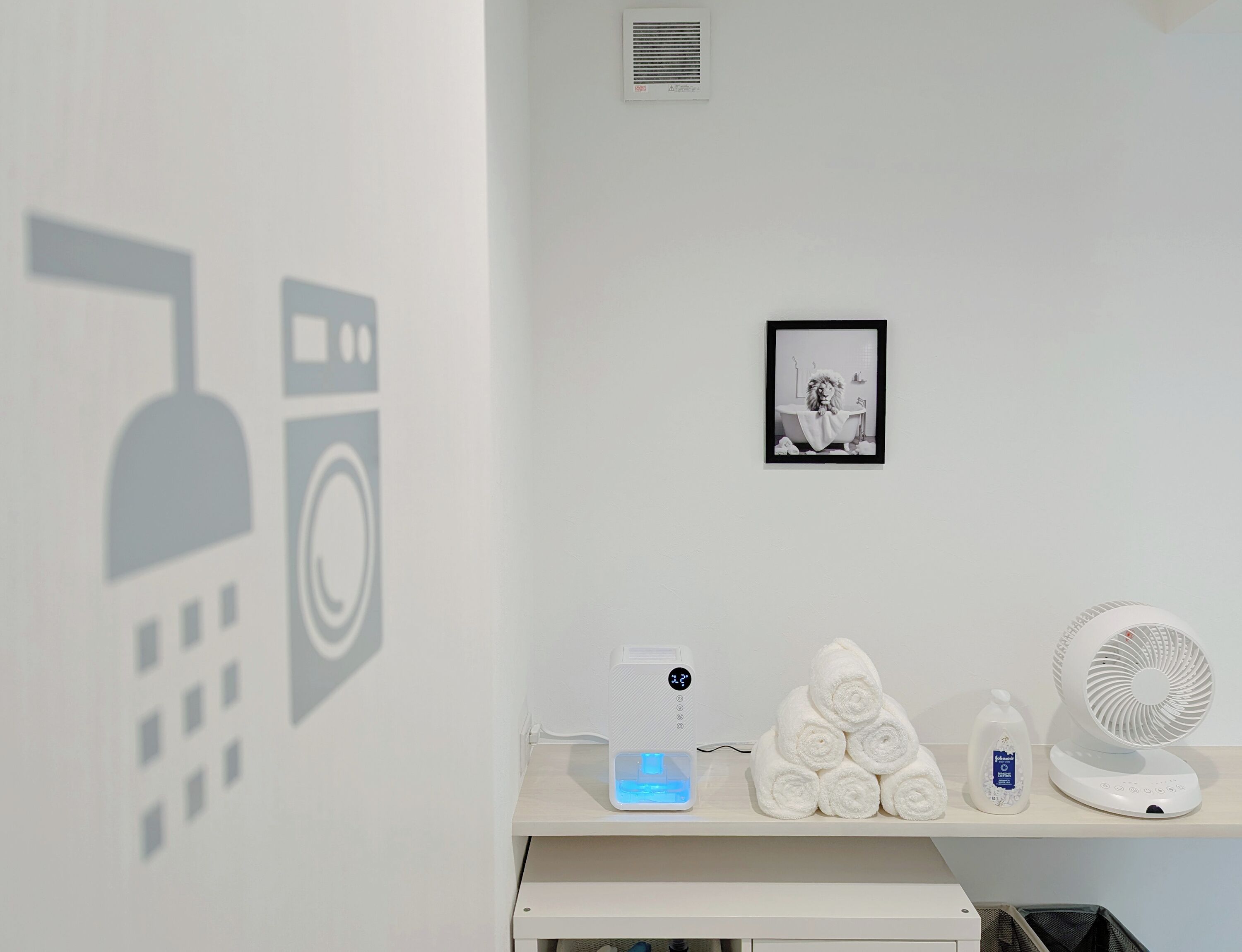 Une salle de bain avec un lavabo blanc, un tableau encadré au mur, un porte-serviettes avec des serviettes, un flacon de savon liquide et un ventilateur blanc.
