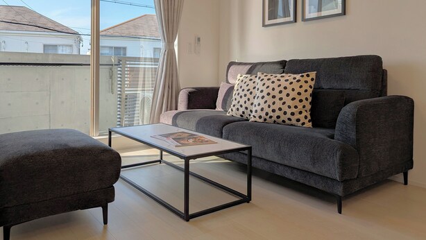 A modern living room with a grey sofa, a matching armchair, and a coffee table. There are two framed pictures on the wall, and a window with curtains. - The FLOW Okinawa (Chatan)