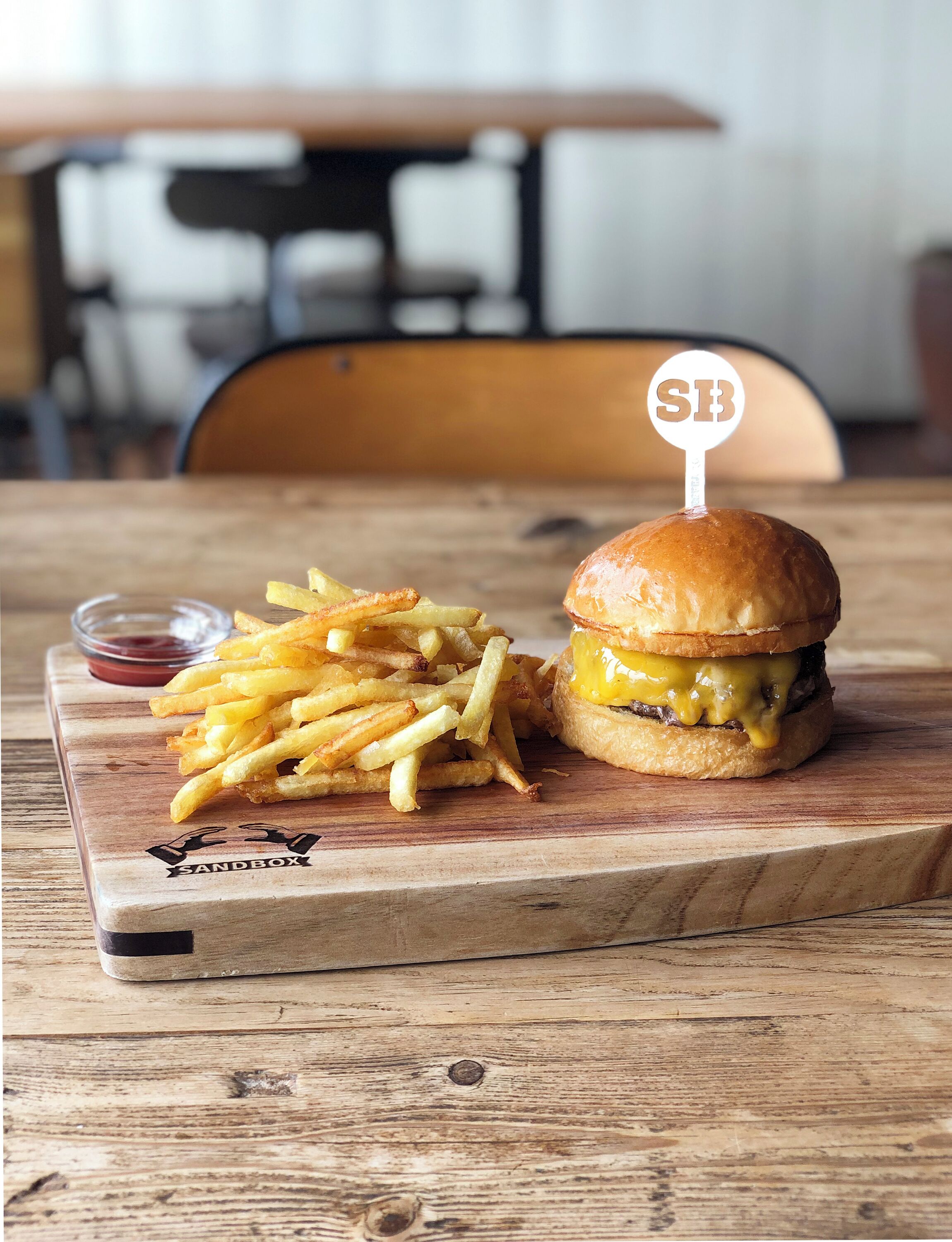 A burger with a sesame seed bun and a 'SB' topper, accompanied by a side of French fries on a wooden board.