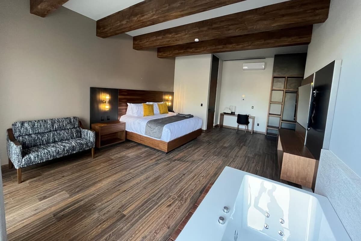 A modern hotel room with a wooden floor, a bed with white linens, a sofa, a bathtub, and a flat-screen TV.