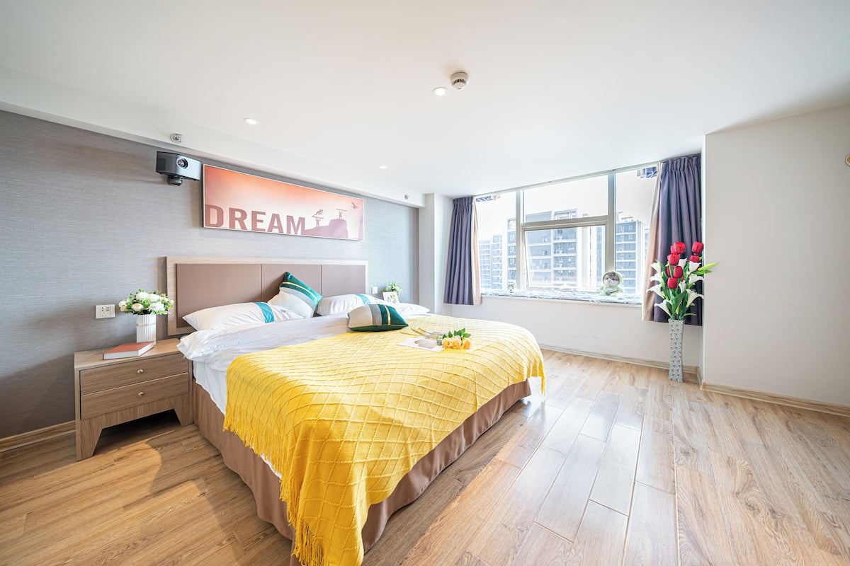 A modern hotel room with a large bed, a nightstand, a wall-mounted air conditioner, a window with curtains, and a city view.