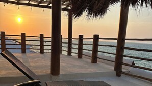 Een zitgedeelte aan het strand met een rieten dak, houten leuningen en uitzicht op de ondergaande zon boven de oceaan.