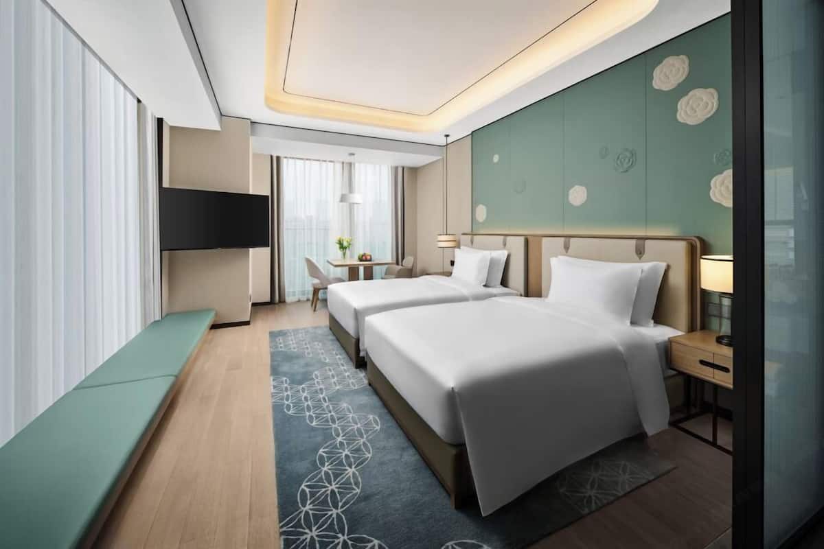 A modern hotel room with two beds, a TV, a desk, and a view of the city.