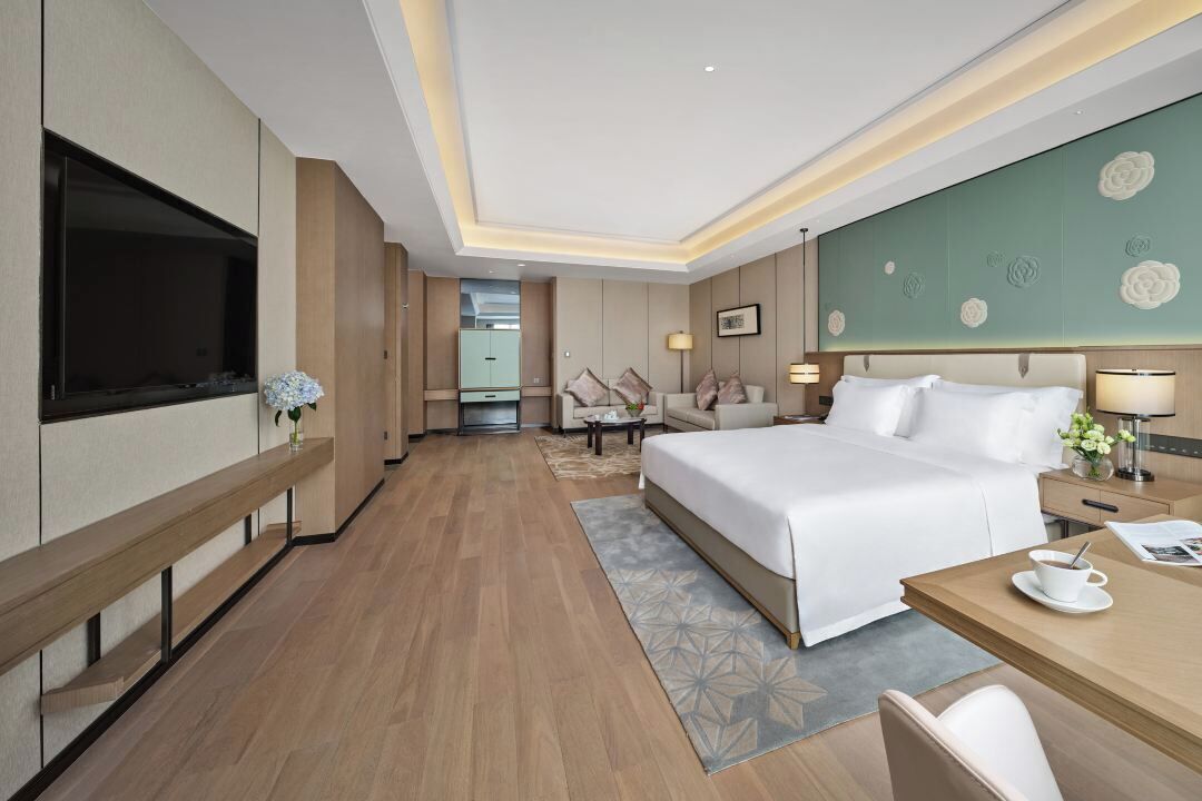 A modern hotel room with a large bed, a desk, a flat-screen TV, and a seating area with a sofa and armchairs.
