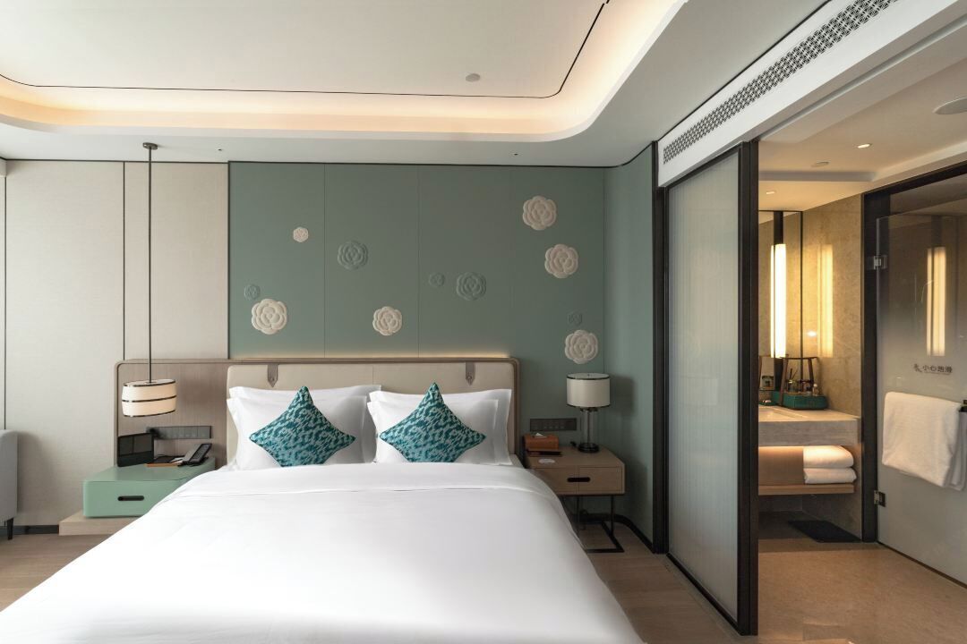 A modern hotel room with a large bed, bedside tables, a nightstand, a mirror, and a bathroom visible through an open door.