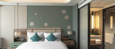 A modern hotel room with a large bed, bedside tables, a nightstand, a mirror, and a bathroom visible through an open door.