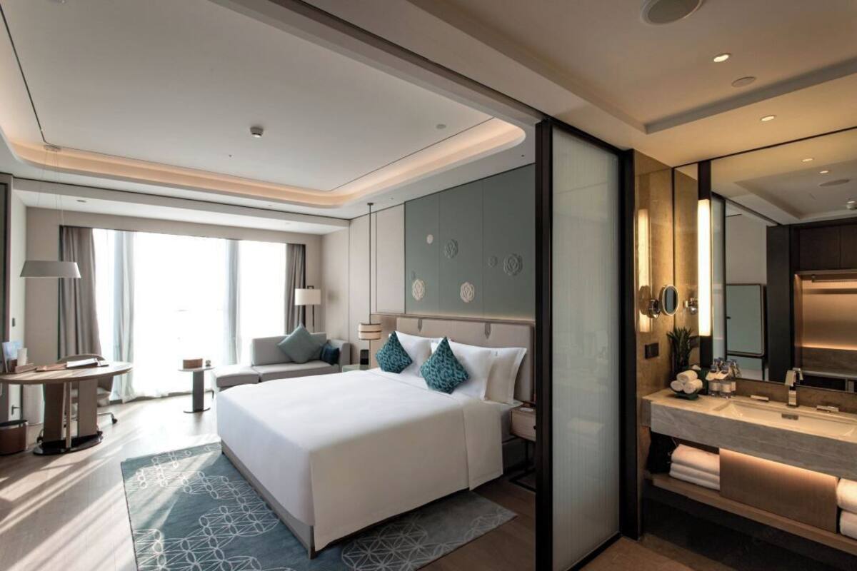 A modern hotel room with a large bed, bedside tables, a desk, and a bathroom with a vanity and mirror.