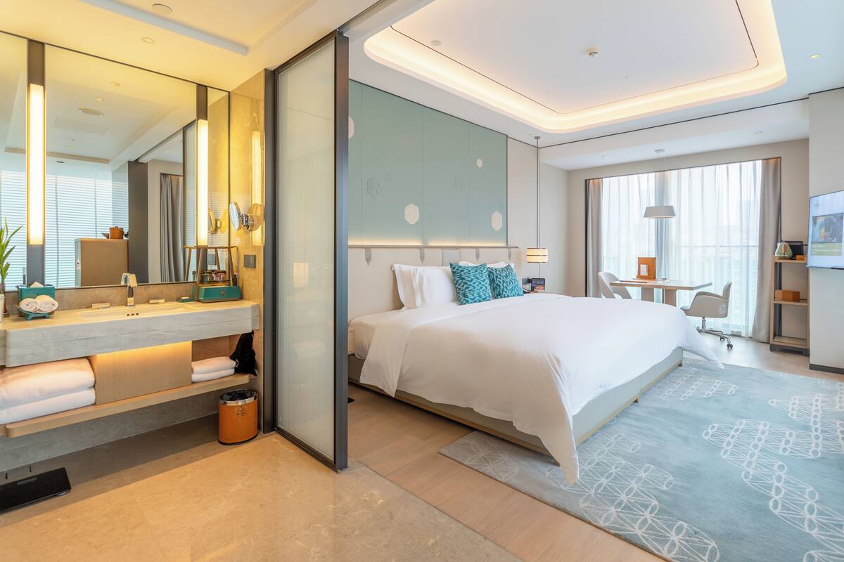 A modern hotel room with a large bed, a bedside table, a mirror, a desk, and a chair.
