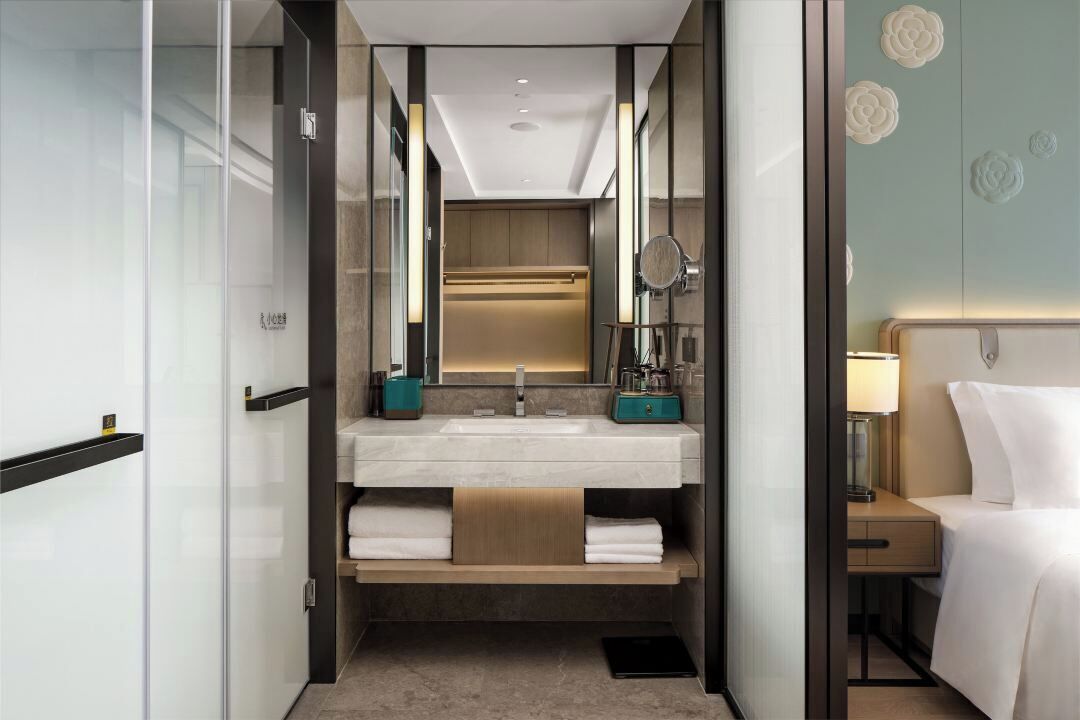A modern bathroom with a glass shower enclosure, a sink with a mirror, and a bedside table with a lamp.