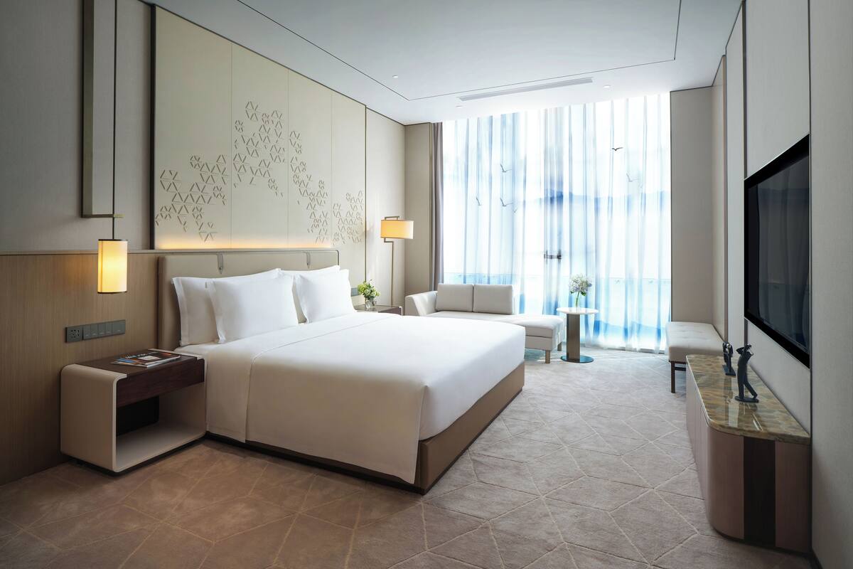 A modern hotel room with a large bed, bedside tables, a TV, and a view of the city through large windows.